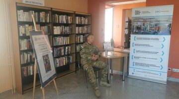 Autor książki w Czytelni Czasopism na tle banerów informacyjnych biblioteki