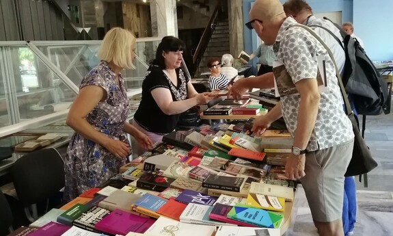 Stół z wydawnictwami, przy nim pracownicy biblioteki a po drugiej stronie uczestnicy wydarzenia.